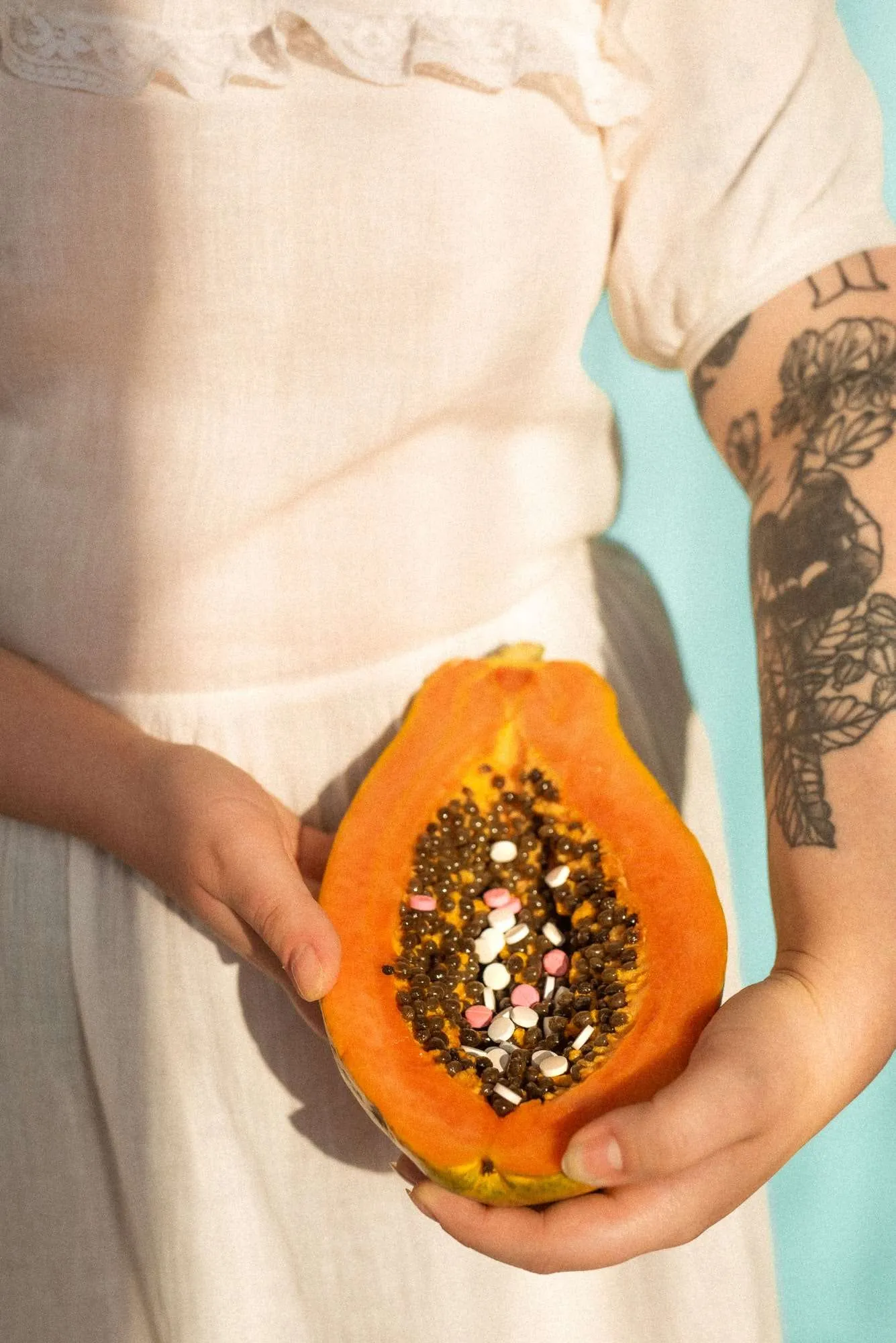 Ręce kobiety trzymające owoc papaji na wysokości swojej talii. Owoc jest przekrojony, a w jego środku znajdują się tabletki.